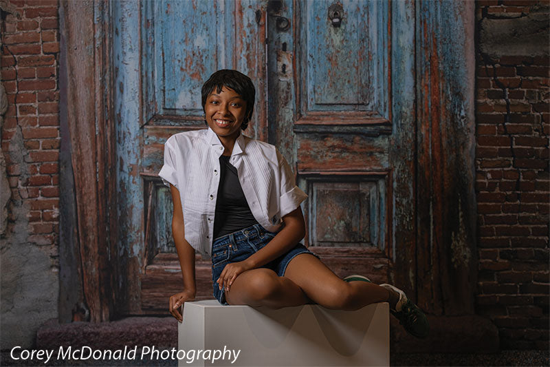 Blue Desire Street, photography backdrop panel for Denny StudioEdge silicone edge frame, shown with a senior girl seated on a white posing cube laughing beside the brick edge SECP-7654