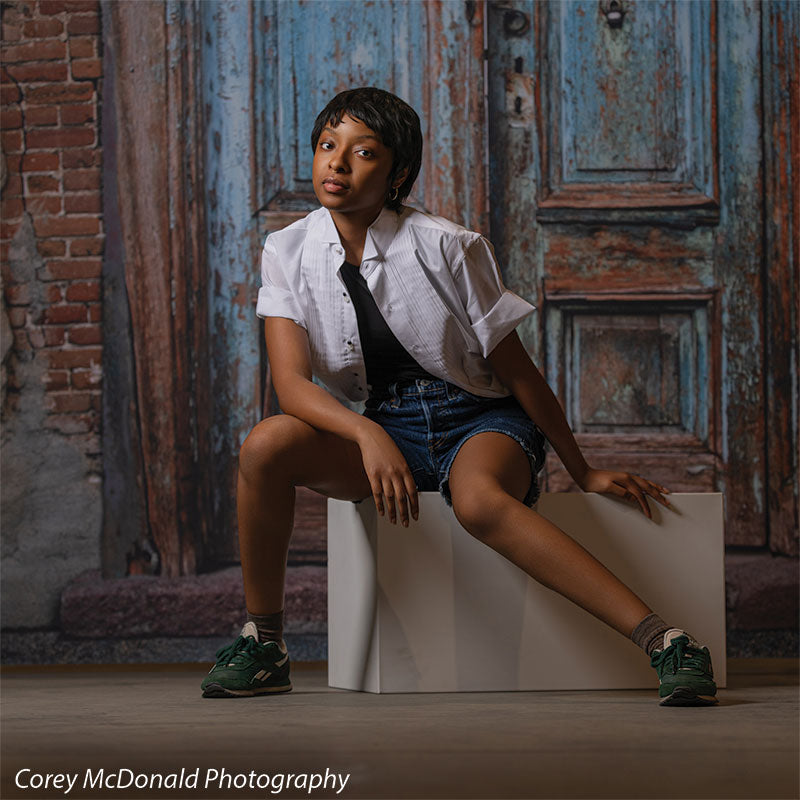 Blue Desire Street, photography backdrop panel for Denny StudioEdge silicone edge frame, shown with a senior girl seated on a white posing cube in front of the weathered door and brick SECP-7654