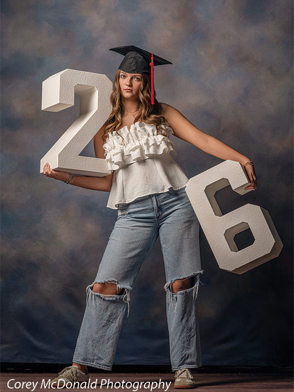 Teen girl with long wavy light brown hair wearing a white ruffled top, ripped jeans, and a graduation cap holds large white numbers two and six while standing in front of a blue gradient portrait backdrop with mottled texture.