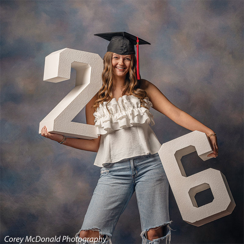 Teen girl with long wavy light brown hair wearing a black graduation cap, white ruffled top, and light blue ripped jeans smiles broadly while holding large white numbers 2 and 6 in front of a blue gray mottled photo backdrop with soft gradient texture.