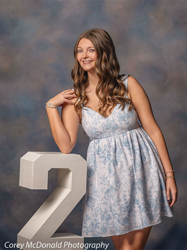 Teen girl with long wavy light brown hair wearing a light blue floral dress smiles while resting her arm on a large white number 2 against a blue gray mottled studio backdrop with soft blended texture.