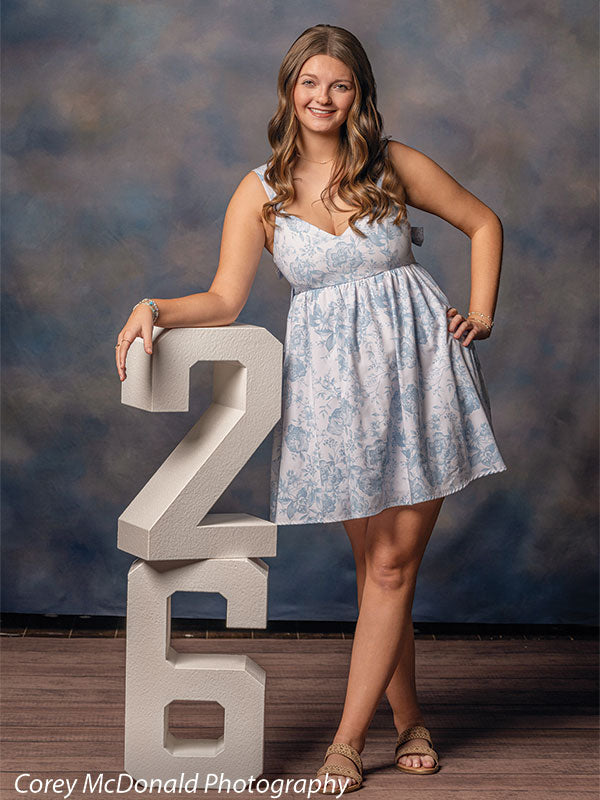 Teen girl with long wavy light brown hair wearing a light blue floral dress smiles while leaning on a large white number 2 with her other hand on her hip in front of a blue gray painted portrait backdrop with mottled effect.