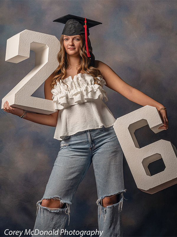 Teen girl with long wavy light brown hair wearing a black graduation cap, white ruffled top, and light blue ripped jeans holds large white numbers 2 and 6 with a neutral expression against a blue gray painted studio backdrop with natural mottled variation.