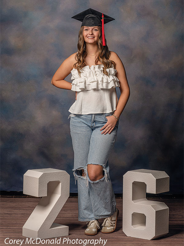 Teen girl with long wavy light brown hair wearing a black graduation cap, white ruffled top, and light blue ripped jeans stands in a relaxed pose smiling between large white numbers 2 and 6 in front of a blue gray painted studio backdrop with soft mottled texture.