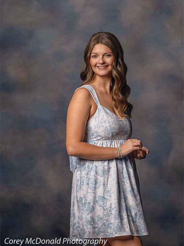 Teen girl with long wavy light brown hair wearing a light blue floral dress turns slightly to the side while smiling, showing bracelets on her wrist against a blue gray portrait backdrop with a soft painterly mottled effect.