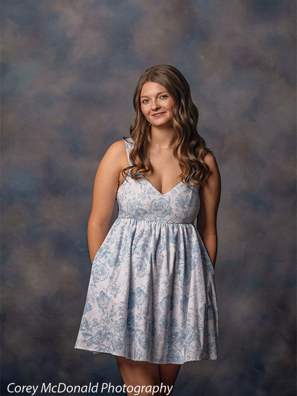 Teen girl with long wavy light brown hair wearing a light blue floral dress smiles softly with hands behind her back in front of a blue gray studio backdrop with smooth mottled portrait texture.