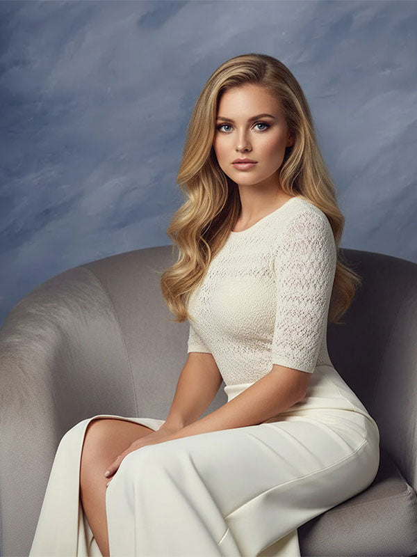 Blonde woman with long wavy hair wearing a white lace top and cream skirt sits on a light gray chair against a blue gray textured photo backdrop.