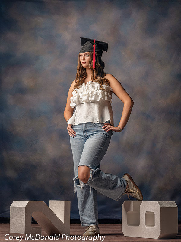 Teen girl with long wavy light brown hair wearing a black graduation cap, white ruffled top, and light blue ripped jeans stands with a wide stance holding large white numbers 2 and 6 in front of a blue gray textured photography backdrop with mottled finish.