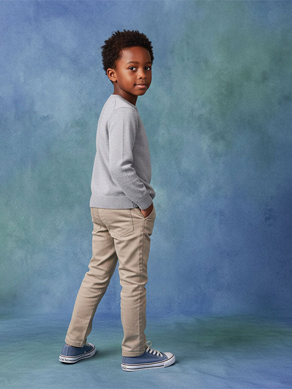 Young boy with short curly hair wearing a gray long sleeve shirt, khaki pants, and blue sneakers stands turned sideways with hands in pockets against a blue green gradient portrait backdrop.