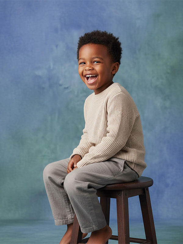 Smiling young boy with short curly hair wearing a beige sweater and light gray pants sits on a wooden stool against a soft blue and green mottled studio backdrop.