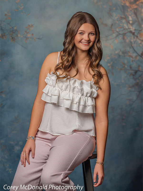 Teen girl with long wavy light brown hair wearing a white ruffled top and pink pants sits on a stool with a relaxed pose and smiling expression against a blue hand painted backdrop with soft foliage accents.