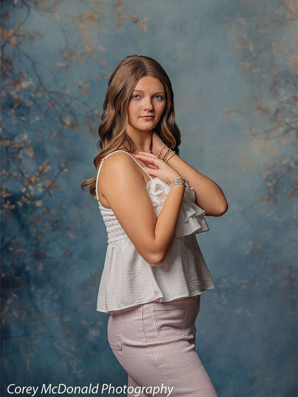 Teen girl with long wavy light brown hair wearing a white ruffled sleeveless top and pink pants holds her hands near her collarbone with a calm expression, posed in front of a blue mottled backdrop featuring soft fall branches.