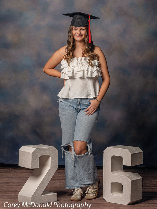 Teen girl with long wavy light brown hair wearing a white ruffled top, jeans, and a graduation cap stands between large white numbers two and six smiling in front of a blue mottled backdrop with subtle gradient texture.