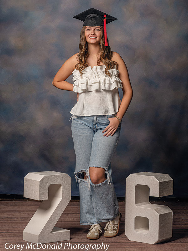 Teen girl with long wavy light brown hair wearing a white ruffled top, ripped jeans, and a graduation cap stands centered and smiling between large white numbers two and six against a blue mottled photography backdrop with soft gradient tones.