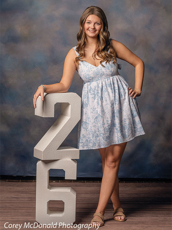 Teen girl with long wavy light brown hair wearing a light blue floral dress leans casually on a large white number two with a relaxed smiling expression in front of a blue mottled portrait backdrop with soft cloud texture.
