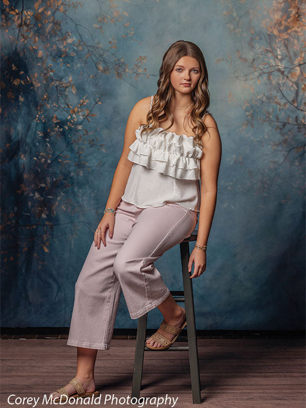 Teen girl with long wavy light brown hair wearing a white ruffled sleeveless top and light pink pants sits on a stool in a relaxed pose in front of a blue painted backdrop with soft autumn branches.