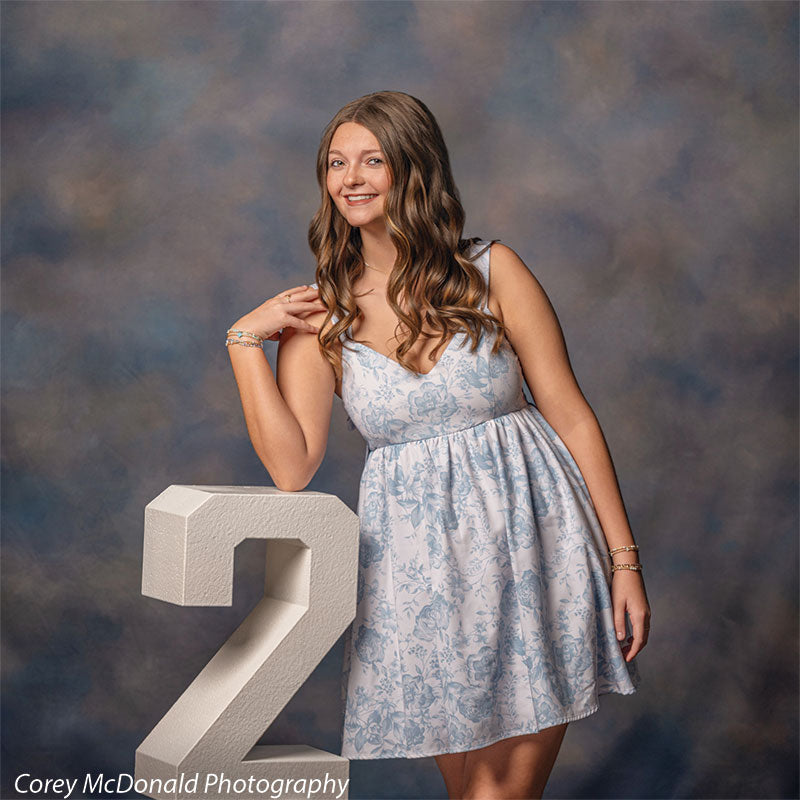 Teen girl with long wavy light brown hair wearing a light blue floral dress leans casually on a large white number two with a relaxed smiling expression in front of a blue portrait backdrop with soft mottled cloud texture.