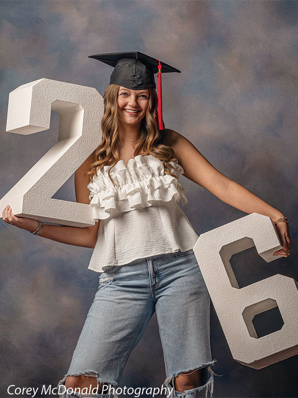 Teen girl with long wavy light brown hair wearing a white ruffled top, ripped jeans, and a graduation cap smiles while holding large white numbers two and six in front of a blue textured portrait backdrop with subtle color blending.