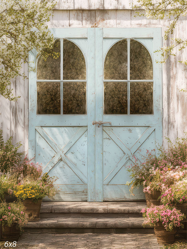 Rustic pale-blue double cottage door framed by stone steps and overflowing spring flower pots in pink, yellow, and white tones. Soft branches and weathered white siding create a dreamy garden entrance feel for portrait photography.