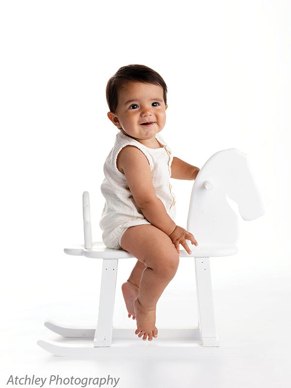 Baby with dark hair in a cream sleeveless romper sitting on a white wooden rocking horse against a bright white seamless photo backdrop, facing forward with a cheerful expression and one hand holding the horse’s head.