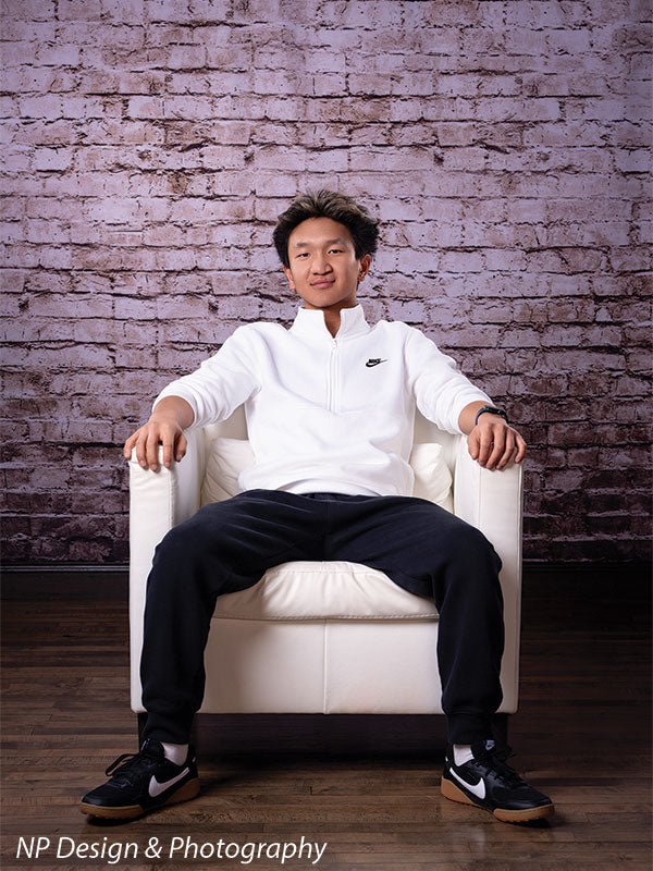 Broadway Brick exposed brick photography backdrop shown with a senior boy in a white quarter-zip seated in a white armchair, full-length CP-7742
