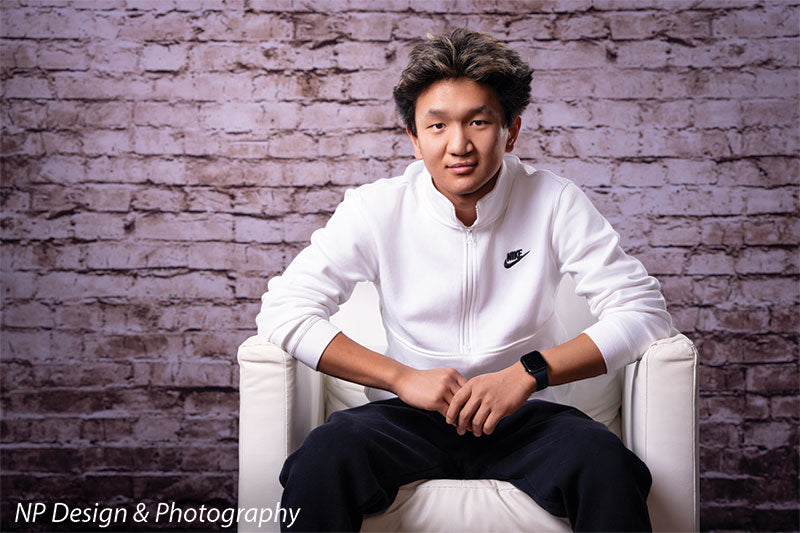 Broadway Brick exposed brick photography backdrop shown with a senior boy seated in a white armchair with hands clasped, wide view CP-7742