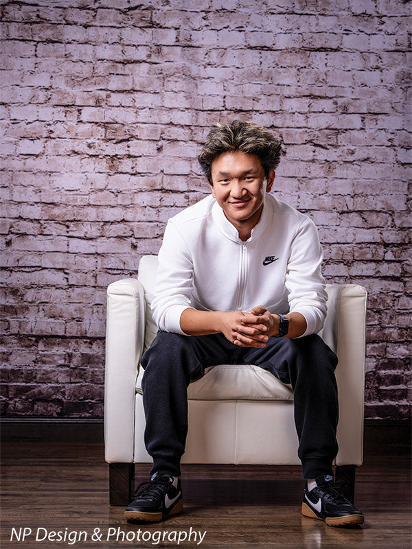 Broadway Brick exposed brick photography backdrop shown with a senior boy seated in a white armchair with hands clasped, smiling CP-7742