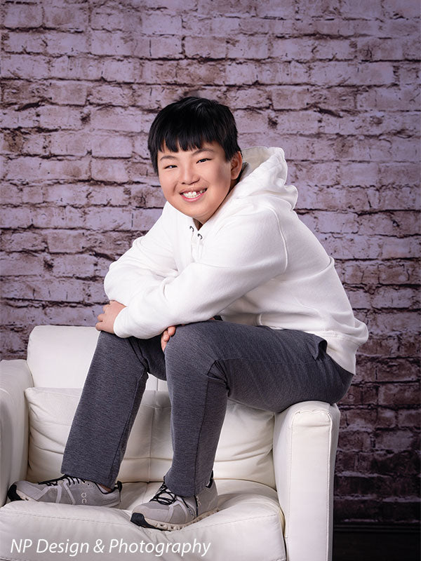 Broadway Brick exposed brick photography backdrop shown with a younger boy in a white hoodie seated in a white armchair with arms crossed CP-7742