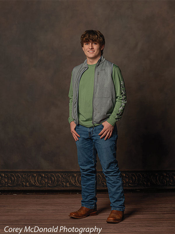 Teen boy with wavy brown hair wearing a green long sleeve shirt, gray vest, blue jeans, and brown boots stands with hands at his sides in front of a brown mottled studio backdrop with soft gradient texture.