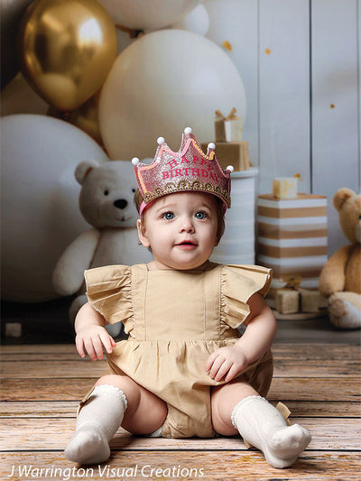 Baby girl wearing a pink 'Happy Birthday' crown surrounded by presents and teddy bears on the Bears and Stars Cake Smash Backdrop.