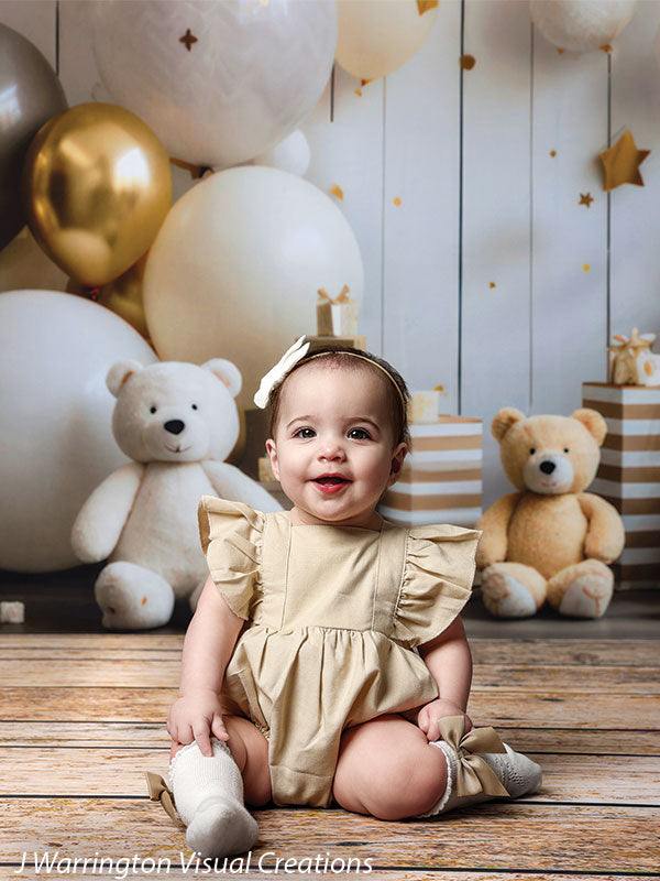 Close-up of a toddler girl sitting in front of white and gold balloons and teddy bears on the Bears and Stars Cake Smash Backdrop.
