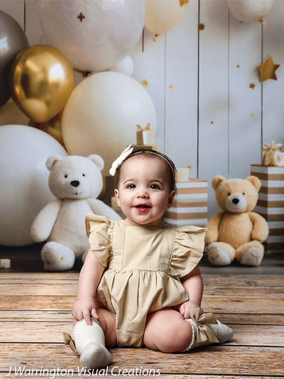 Close-up of a toddler girl sitting in front of white and gold balloons and teddy bears on the Bears and Stars Cake Smash Backdrop.
