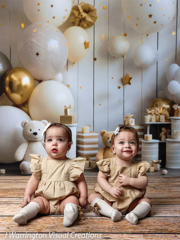 Two toddlers sitting on rustic wood flooring surrounded by festive white and gold balloons, teddy bears, and gifts in the Bears and Stars Cake Smash Backdrop.