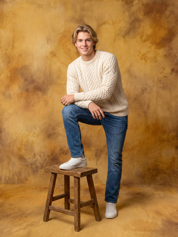Caramel Drift caramel gold mottled texture photography backdrop, shown with a young man in a cream cable-knit sweater and jeans seated on a wood stool CP-9021