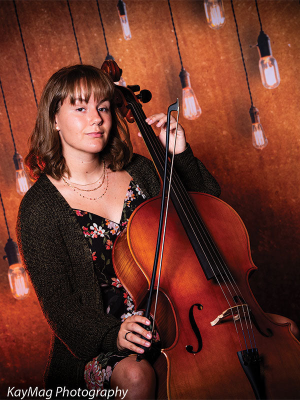 Female student musician posing with a cello in front of a rustic backdrop with string lights, ideal for artistic senior portraits with an instrumental theme.