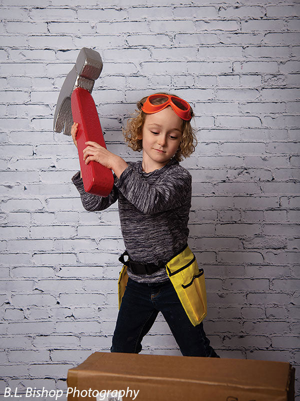 Smiling child wearing a red toy miner’s helmet and goggles poses with a giant hammer prop against a white brick backdrop in a styled studio session.