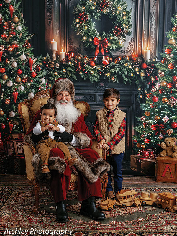 Santa Claus seated in an ornate chair holding a baby in brown overalls while a young boy in a red plaid shirt and tan vest stands beside him, with wooden toy trains on the floor and a decorated Christmas tree, wreath, garland, candles, and fireplace in the festive holiday backdrop.