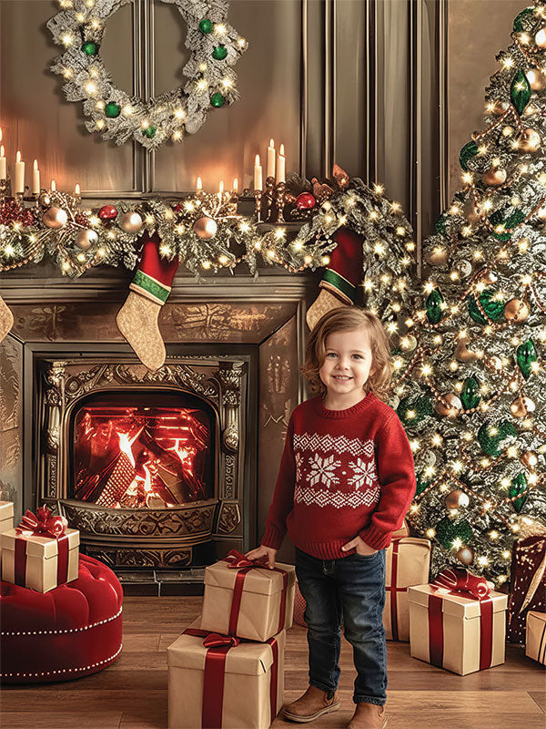 A young child wearing a red sweater stands among wrapped gifts near a lit fireplace and decorated Christmas tree, posed in front of a festive holiday backdrop with garlands, candles, stockings, and warm glowing lights.

