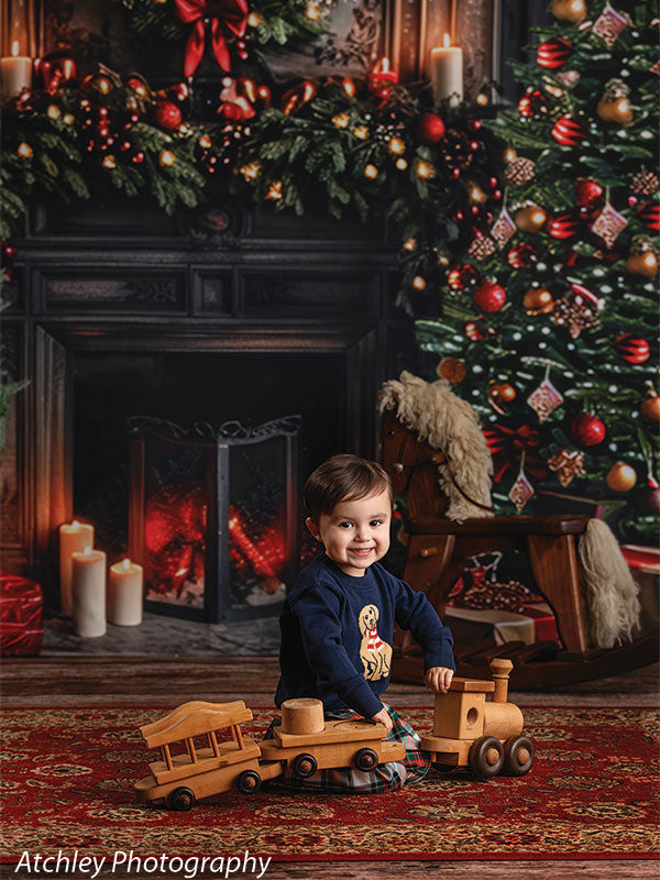 A smiling toddler sits on a patterned rug playing with wooden toy trains, posed in front of a cozy Christmas backdrop featuring a decorated tree, fireplace, candles, and holiday garland.
