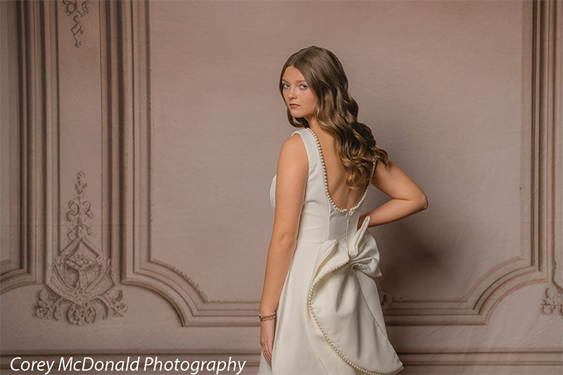 Teen girl with long wavy light brown hair wearing a sleeveless white dress stands turned away looking over her shoulder, showing the bow detail on the back of the dress against a classic beige wall backdrop with ornate panel accents.