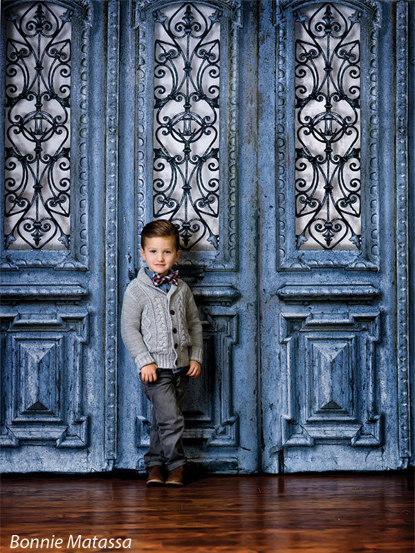 Young boy in a cable-knit sweater and dress pants poses in front of a distressed blue vintage door with ornamental ironwork, ideal for formal child portraits.