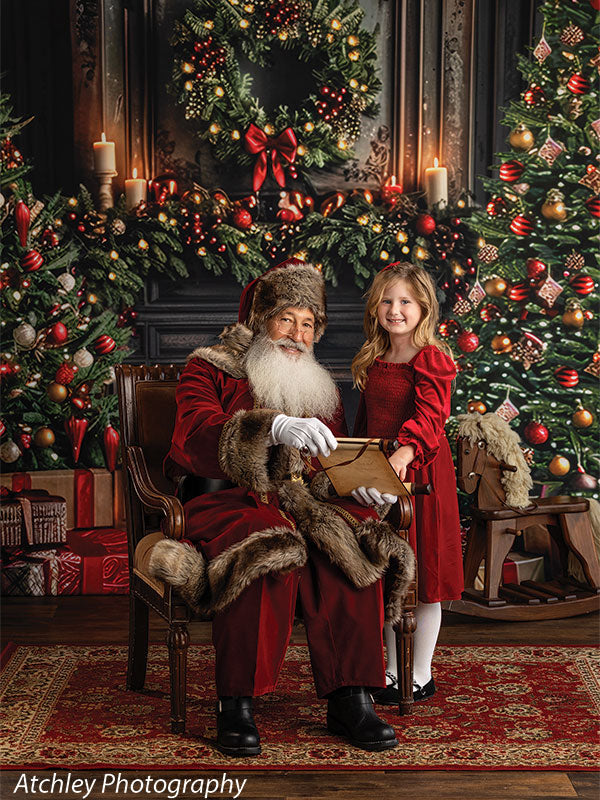 Santa Claus seated in a vintage armchair beside a smiling young girl in a red dress, with a decorated Christmas tree, wreath, garland, candles, wrapped presents, and a lit fireplace creating a classic holiday living room backdrop.