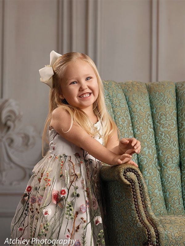 A young girl wearing a floral embroidered dress smiles while resting her arms on a green upholstered chair, posed against a classic elegant wall backdrop with subtle floral accents.
