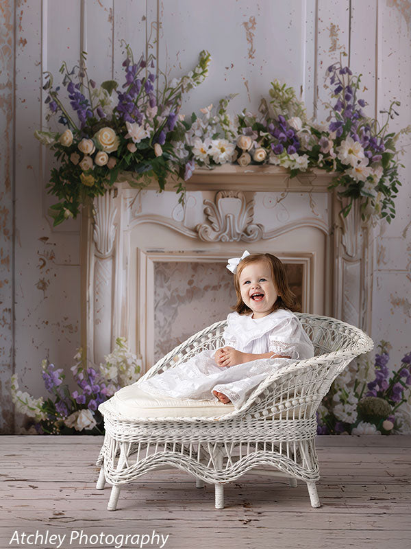 Toddler girl in a white dress and bow, posed on a white wicker chaise lounge with wood floor, against an ornate vintage floral mantle backdrop with cascading flowers.