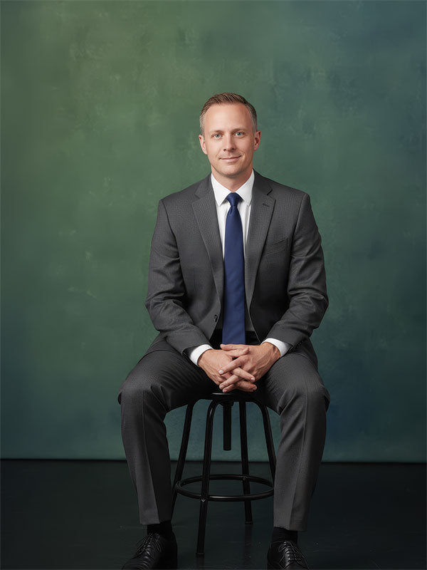 A man in a gray suit and blue tie sits on a stool with hands folded, posed against a classic green painted studio backdrop with soft texture and a clean professional look.
