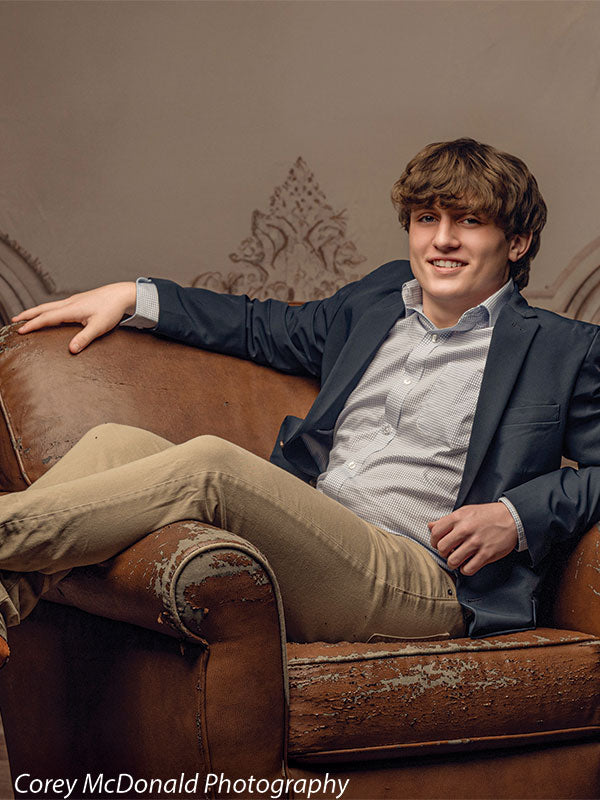 Teen boy with wavy light brown hair wearing a blue blazer and khaki pants reclines in a distressed brown leather armchair smiling slightly, positioned in front of a classic beige ornate wall backdrop with aged panel details.
