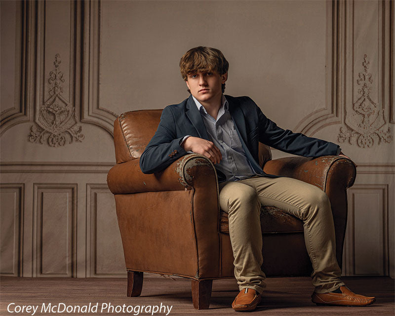 Teen boy wearing a navy blazer, light blue shirt, khaki pants, and brown shoes sits relaxed in a brown leather armchair in front of a beige ornate wall panel backdrop with elegant decorative molding.