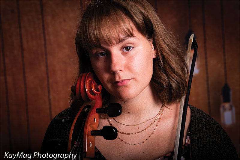 Close-up of a young female musician holding a cello bow in front of a moody wood plank backdrop with hanging Edison bulbs, great for expressive senior or music-themed photography.