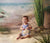 Portrait of baby on studio sand setup with painted beach and waves backdrop, ideal for summer-themed or milestone sessions.