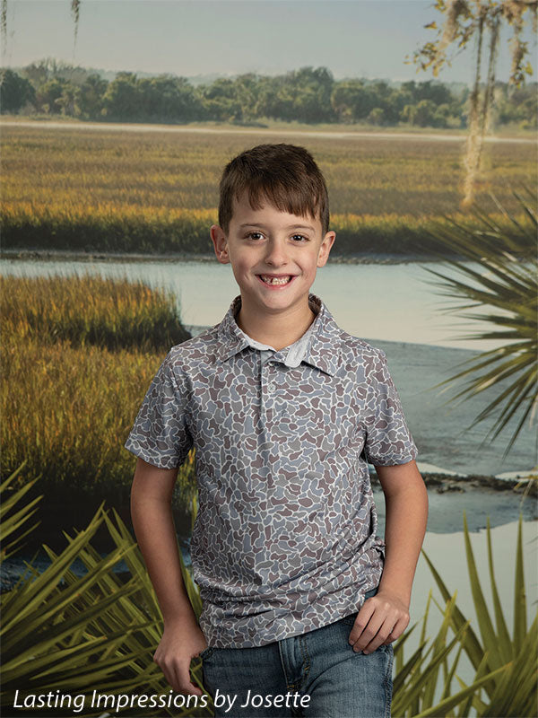 Coastal Marsh View photography backdrop showing golden marsh grass and a tidal creek with a distant treeline and palmetto fronds, shown with a young boy in a gray patterned polo with a hand in his pocket CP-8486
